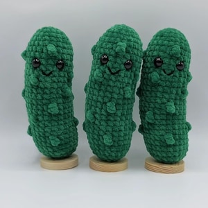 May include: Three handmade, green crocheted pickle plushies with smiling faces and small wooden bases. The pickles have black button eyes and small, textured bumps. The plushies are displayed against a white background.