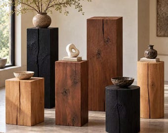 May include: A collection of wooden pedestals and stools in various heights and finishes, including natural light oak, medium brown, and charred black wood. Each piece features a vertical split and a solid, block-like or cylindrical form.