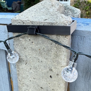 May include: A black bracket secures a string of clear globe lights to a concrete post. The lights are strung on a green wire, and the bracket is attached with a black strap. A blue painted wooden fence is in the background.