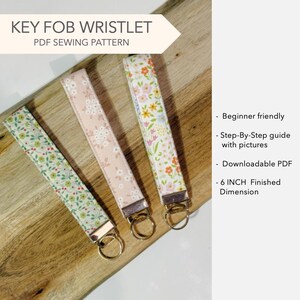 May include: Three fabric key fob wristlets with floral patterns in green, pink, and multi-colored designs. Each wristlet features a silver-tone metal hardware clasp and a split ring. The text reads KEY FOB WRISTLET PDF SEWING PATTERN and 6 INCH Finished Dimension.