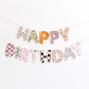 May include: A Happy Birthday banner featuring individual fabric letters strung together. Each letter displays a unique pattern, including gingham, floral, stripes, and solid colors in shades of pink, orange, brown, green, and white with scalloped edges.