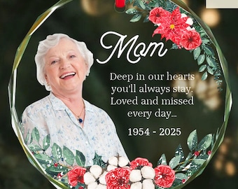 May include: A clear, faceted glass ornament with a photo of a smiling woman, surrounded by floral accents and the word "Mom." Text reads "Deep in our hearts you'll always stay. Loved and missed every day... 1954-2025."