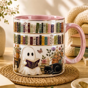 May include: A white ceramic mug with a pink interior and handle, featuring a wrap-around illustration of two shelves of colorful books, a ghost reading a purple book, a black cat, potted flowers, a candle, and a small butterfly.