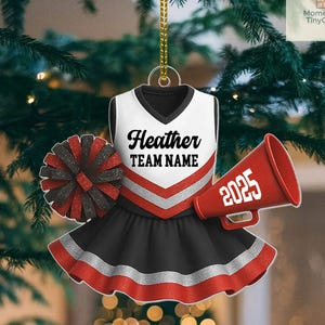 May include: A cheerleader-themed ornament featuring a white, black, and red uniform with the name "Heather" and "TEAM NAME". Includes a red megaphone with "2025" and a pom-pom. The ornament hangs from a gold chain.