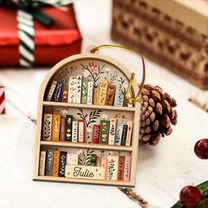 May include: Wooden bookshelf ornament with a gold hanging string. The ornament features a miniature bookshelf design with painted books and floral accents. The name "Julie" is written on a small wooden sign. The ornament is a charming decorative piece.