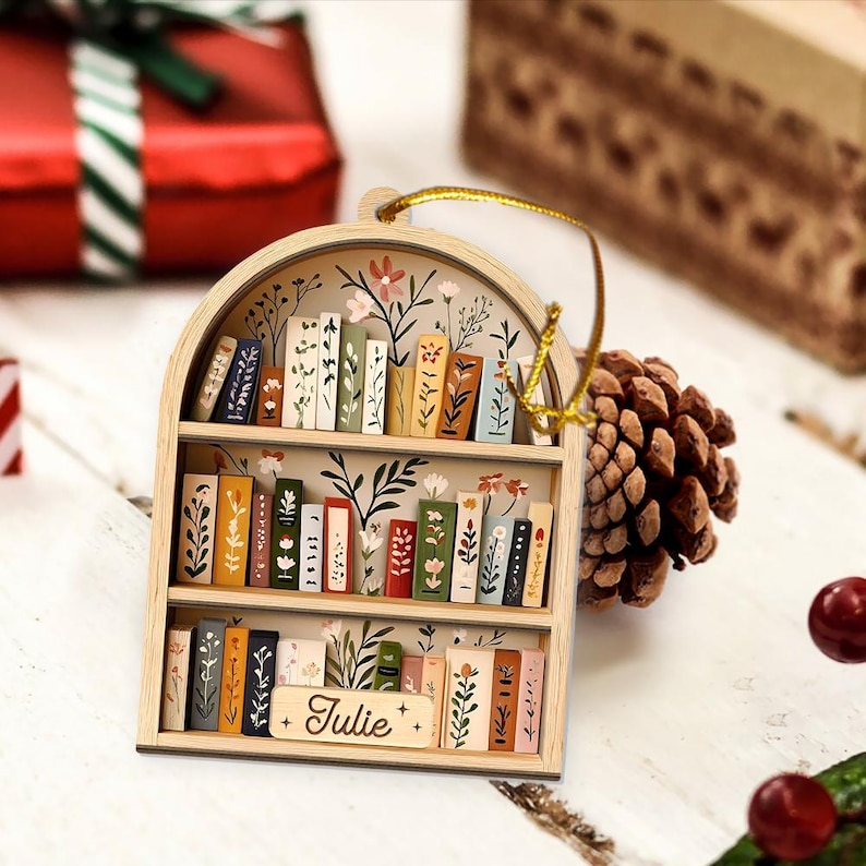 May include: Wooden bookshelf ornament with a gold hanging string. The ornament features a miniature bookshelf design with painted books and floral accents. The name "Julie" is written on a small wooden sign. The ornament is a charming decorative piece.