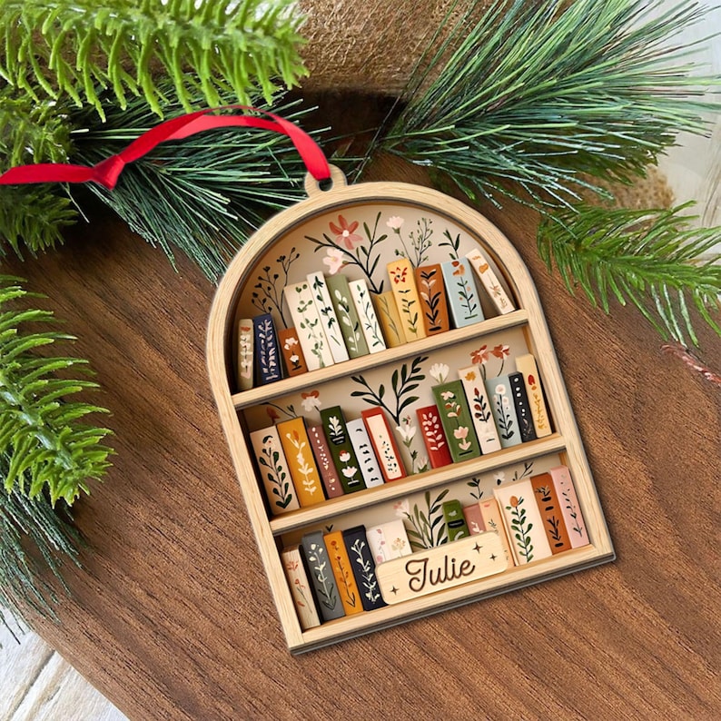May include: Wooden bookshelf ornament with a red ribbon for hanging. The ornament features a miniature bookshelf design with painted books and floral accents. The name "Julie" is written on a small sign.