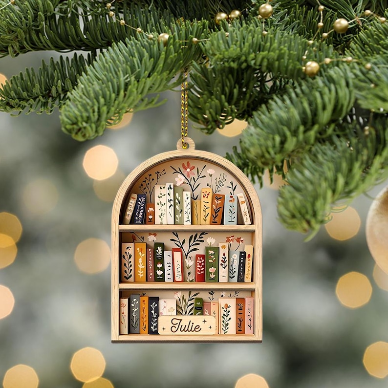 May include: Wooden bookshelf ornament with a gold chain for hanging. The ornament features a miniature bookshelf design with painted books and floral accents. The name "Julie" is written on a small sign at the bottom. The ornament is hanging on a Christmas tree.