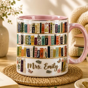 May include: A white ceramic mug with a pink interior and handle, decorated with colorful book spines arranged in rows. The mug is personalized with the name "Mrs. Emily" in gold script, with floral and butterfly accents.