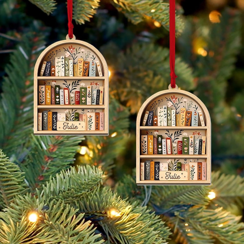 May include: Two wooden bookshelf ornaments with red ribbons for hanging. Each ornament features a miniature bookshelf filled with colorful books and floral accents. The name "Julie" is inscribed on a small wooden plaque on each shelf.