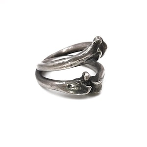Könnte beinhalten: Ein silberfarbener Ring mit einem einzigartigen, knochenartigen Design. Der Ring hat zwei ineinandergreifende Bänder mit strukturierten Details und einem leicht oxidierten Finish. Das Design verleiht dem Ring eine rustikale, organische Ästhetik.