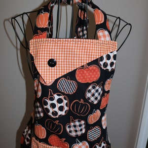 May include: Black and orange Halloween apron with a pumpkin print. The apron has a gingham pocket and a gingham trim.