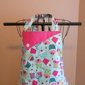 May include: A light blue apron with a pink trim and a white button. The apron is covered in a pattern of colorful cupcakes.