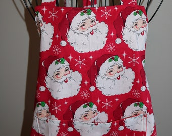 May include: Red Christmas apron with a repeating pattern of Santa Claus faces and snowflakes. The apron has a large pocket in the front.