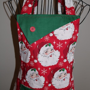 May include: A red and green Christmas apron with a Santa Claus print. The apron has a green pocket with a red button and a green bib with a red button.