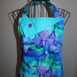 May include: A colourful apron with a vibrant floral pattern in shades of blue, purple, and green. The apron features a turquoise pocket and accents, with a matching button detail. The apron is displayed on a black metal form.