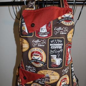 May include: A red and brown cotton apron with a coffee theme. The apron has a large pocket and adjustable straps. The fabric features a variety of coffee-themed designs, including coffee cups, coffee beans, and coffee grinders. The text on the apron includes "Coffee Time", "House Blend", "Premium Quality Coffee", and "Hot & Fresh Morning Coffee".