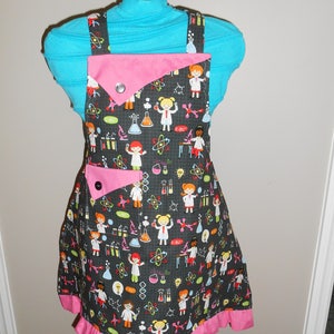 May include: A black and pink apron with a science theme. The apron features a pattern of girls in lab coats, beakers, and other science-related imagery. The apron has a pink ruffle around the bottom.