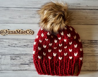 May include: Red knit beanie with a white heart pattern and a brown faux fur pom pom. The beanie has a small brown leather tag with the text "@JunaMaeCo" on it.
