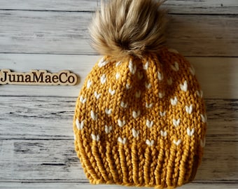 May include: A mustard yellow knit beanie with a white heart pattern and a brown faux fur pom pom. The beanie is on a white wood background with a wooden sign that says "@JunaMaeCo".