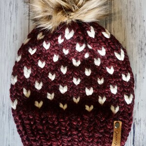 May include: A burgundy knit beanie with a white heart pattern and a faux fur pom pom. The beanie has a brown leather tag with the text "Juna Mae Co."