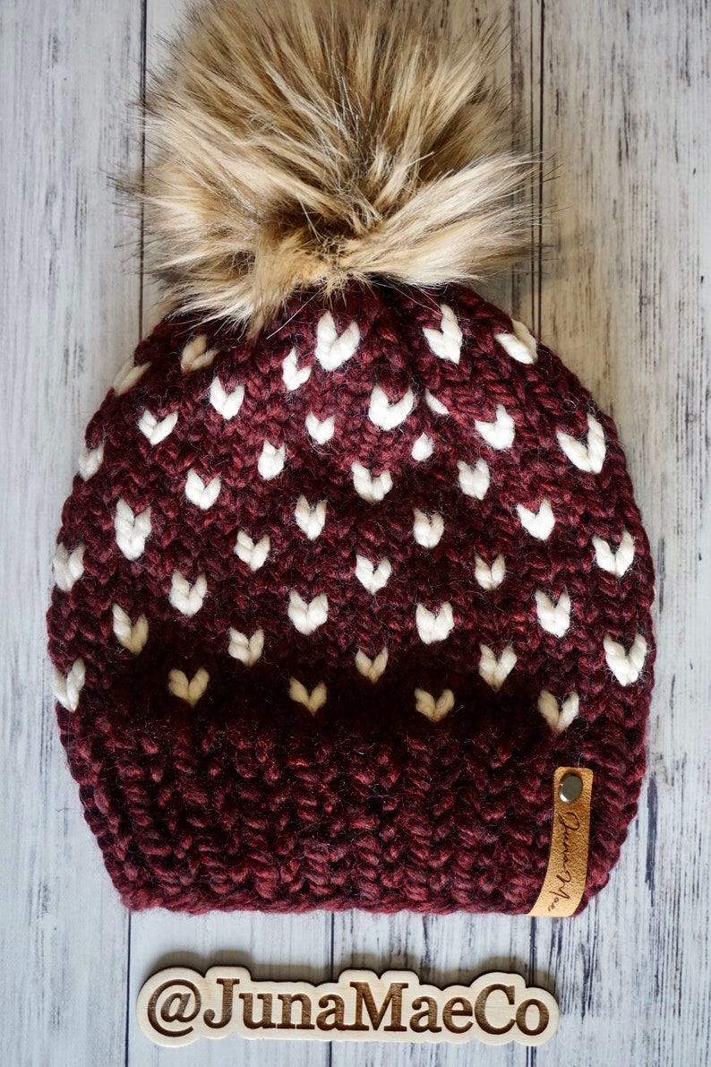 May include: A burgundy knit beanie with a white heart pattern and a faux fur pom pom. The beanie has a brown leather tag with the text "Juna Mae Co."