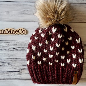May include: A burgundy knit beanie with a white heart pattern and a faux fur pom pom. The beanie has a brown leather tag with the text "@JunaMaeCo" on it.