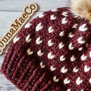 May include: A maroon knitted beanie with a white heart pattern and a brown faux fur pom pom. The beanie has the text "@JunaMaeCo" on a wooden tag.