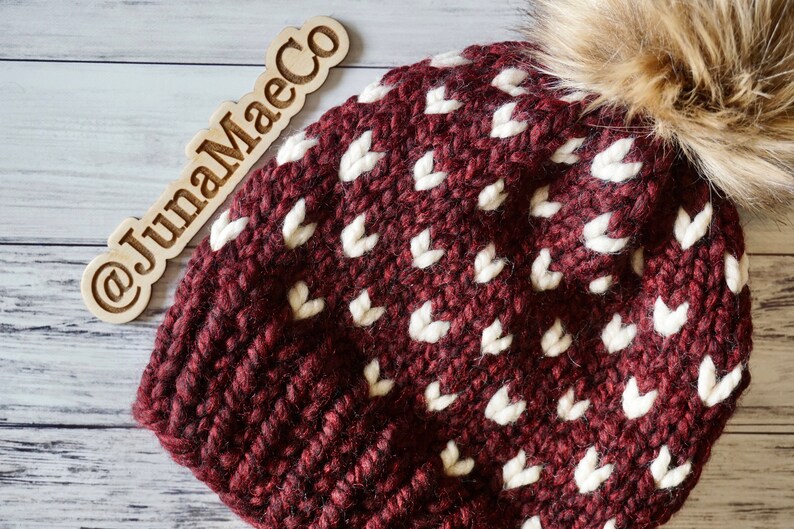 May include: A maroon knitted beanie with a white heart pattern and a brown faux fur pom pom. The beanie has the text "@JunaMaeCo" on a wooden tag.