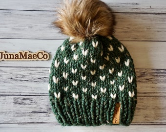 May include: A green knit beanie with a white heart pattern and a brown faux fur pom pom. The beanie has a brown leather tag with the text "Juna Mae Co."