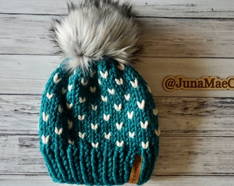 May include: Teal knitted beanie with a white heart pattern and a white faux fur pom pom. The beanie has a brown leather tag with the text "@JunaMaeCo" on it.