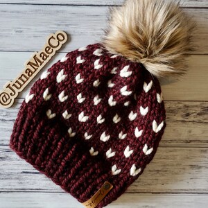 May include: A maroon knitted beanie with a white heart pattern and a brown faux fur pom pom. The beanie has a brown leather tag with the text "JunaMaeCo" on it.