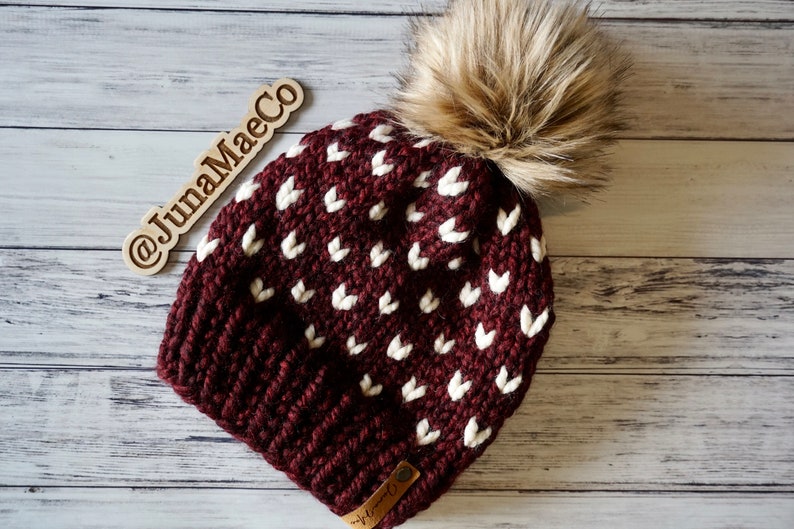 May include: A maroon knitted beanie with a white heart pattern and a brown faux fur pom pom. The beanie has a brown leather tag with the text "JunaMaeCo" on it.