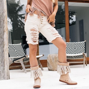 May include: A woman wearing a light brown sleeveless top with fringe detailing, paired with distressed white denim shorts. She is also wearing beige boots with fringe accents. The background features a white wall with the text "Chintamani Alchemy".