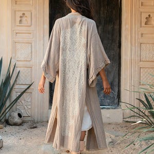 May include: A long, beige, open-front kimono-style robe featuring wide sleeves and a textured, cream-colored lace panel running down the center of the back. The garment is worn over a white base layer and reaches mid-calf length with side slits.
