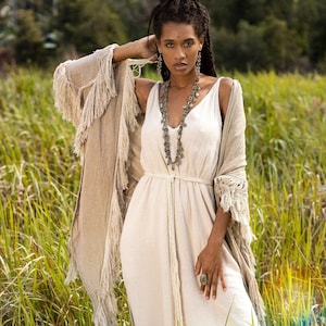 May include: A woman in a cream dress and fringed shawl poses in a field. The dress has a V-neck and a rope belt. The shawl is a light brown colour. The image includes the text "chintamani alchemy".