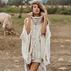 May include: A woman in a cream-coloured, fringed poncho with a high collar and button closure, paired with shorts. The background shows a field, palm trees, and a cow grazing. The text "@hintamanl ALCHEMY" is in the upper left corner.