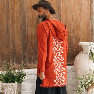 May include: A long-sleeved, hooded, burnt-orange tunic featuring a white geometric triangle pattern down the back and a fringed hem, paired with loose-fitting black trousers. The text "chintamani ALCHEMISTRY" is visible in the upper left corner.