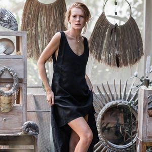 May include: A woman wearing a black sleeveless dress. The dress has a low neckline and a high-low hemline. The background includes wooden shelves with decorative items, a rug, and hanging circular decorations.