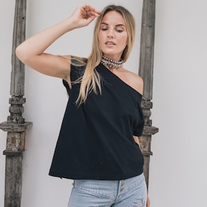 May include: A black, one-shoulder top paired with light blue, distressed jeans. The model is wearing a silver choker necklace. The background features weathered wooden architectural elements against a white wall. The overall aesthetic is casual and bohemian.