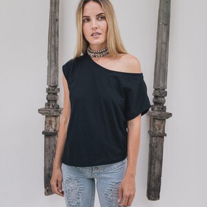 May include: Black off-the-shoulder top with a single short sleeve. The top is paired with light blue distressed jeans. A silver choker necklace is worn. The background features two weathered wooden pillars and a white wall.