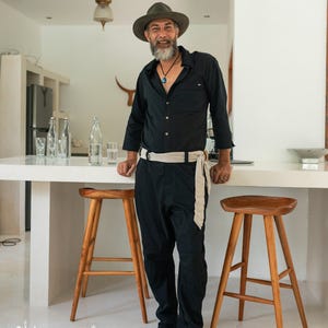 May include: A black long-sleeved button-down shirt and matching black trousers with a light beige fabric belt tied at the waist. The outfit is paired with a dark olive green fedora hat and a blue pendant necklace on a black cord.