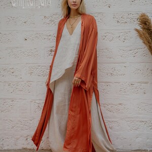 May include: A woman in a rust-coloured long open front robe over a white top and light beige trousers. The robe has long sleeves and a flowing design. The outfit is paired with tan platform shoes. The text "chintamani" is visible in the upper left corner.