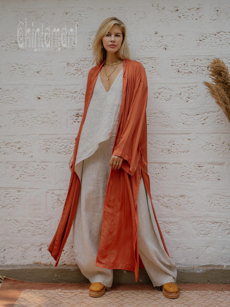 May include: A woman in a rust-coloured long open front robe over a white top and light beige trousers. The robe has long sleeves and a flowing design. The outfit is paired with tan platform shoes. The text "chintamani" is visible in the upper left corner.