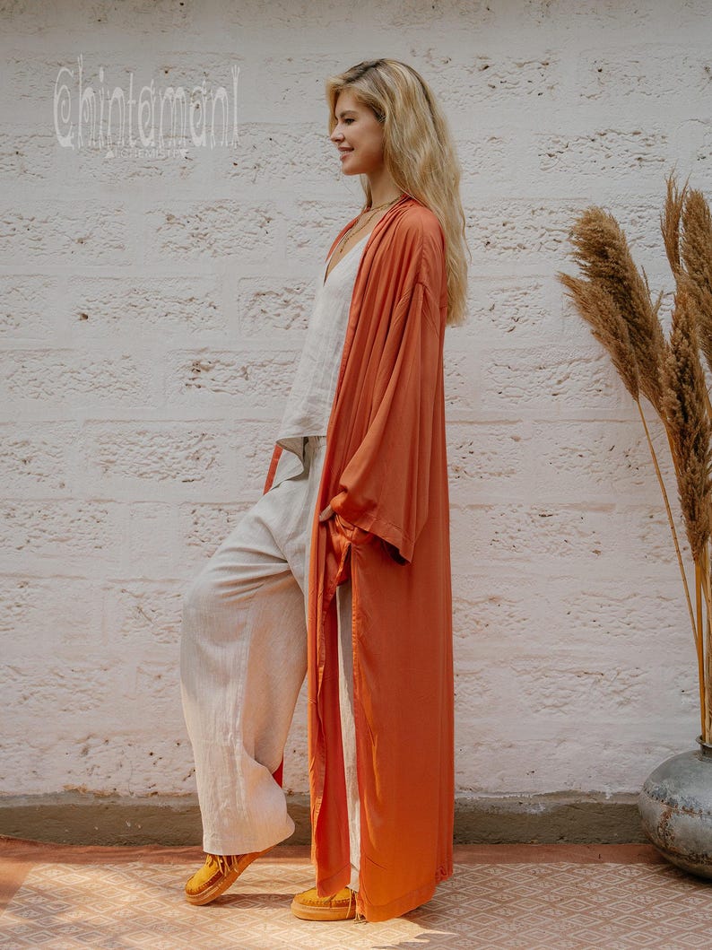 May include: A woman in a rust-coloured long cardigan, white top, and light-coloured trousers, paired with yellow shoes. The background includes a white brick wall and dried plants in a vase. The text "chintamani" is visible.