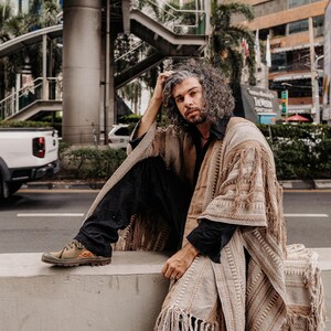 Puede incluir: Un poncho beige abierto por delante con intrincados patrones tejidos y flecos largos en los bordes. La prenda se lleva sobre una camisa negra de cuello y pantalones negros, combinada con zapatillas bajas de color verde oliva y naranja.