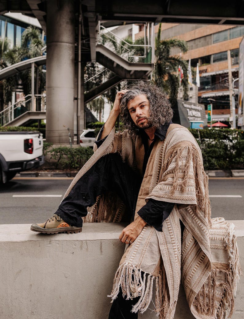 Puede incluir: Un poncho beige abierto por delante con intrincados patrones tejidos y flecos largos en los bordes. La prenda se lleva sobre una camisa negra de cuello y pantalones negros, combinada con zapatillas bajas de color verde oliva y naranja.