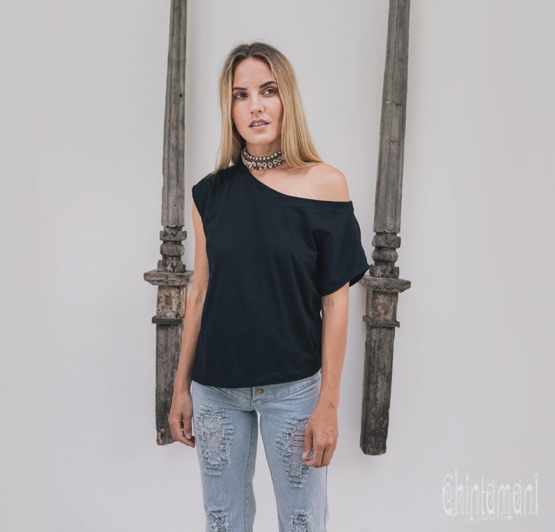 May include: Black off-the-shoulder top with a single short sleeve. The top is paired with light blue distressed jeans. A silver choker necklace is worn. The background features two weathered wooden pillars and a white wall.