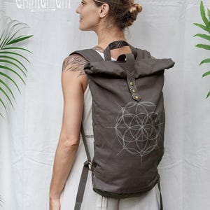 May include: A dark grey roll-top backpack featuring a white geometric flower of life pattern on the front. The bag includes a top handle, adjustable shoulder straps, and a central strap secured with two brass-coloured metal snaps. Text reads Chintamani.