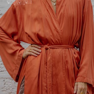 May include: A burnt orange, long-sleeved robe with a wrap-around design and a matching belt. The robe has a silky texture and features subtle vertical stitching details. The sleeves are wide and flowing. The brand name "chintamani" is visible.